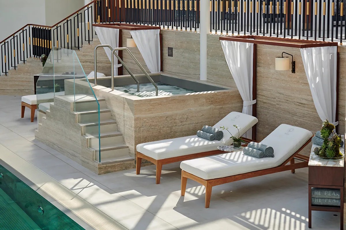 An indoor pool with white reclining loungers, marble pavements and wall, a sea green-pool, and wooden furniture.