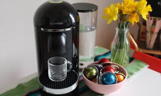 En Nespresso Vertuo Plus står på ett bord bredvid en skål med kaffekapslar, redo att brygga en kopp kaffe.
