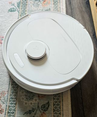 A white robot vacuum on top of a white and green patterned floral rug, dark wooden floorboards, and a light wooden table