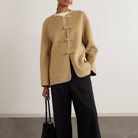 A model wearing a beige wool jacket, black trousers and holding a black handbag.