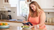 Woman sits at a counter writing in a journal with a plate of food and glass of water in front of her