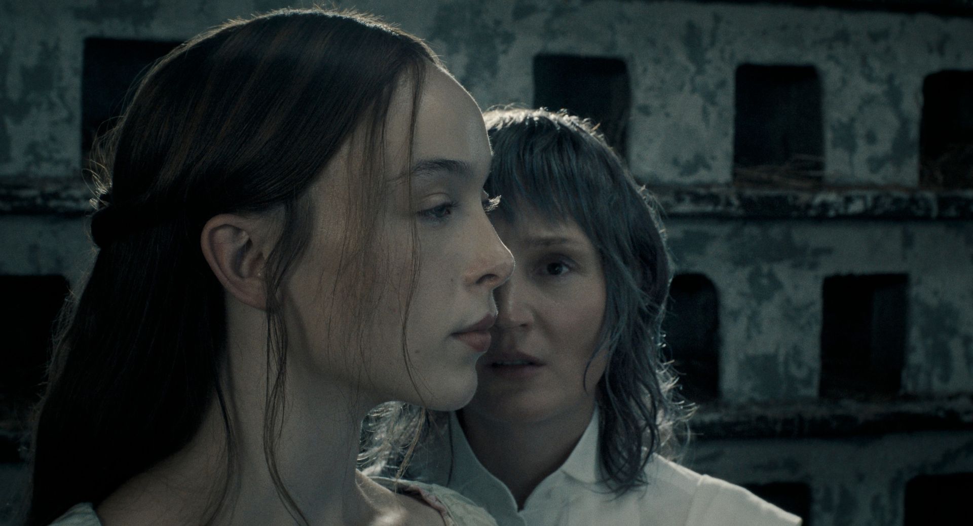 ella beatty as lizzie borden looking ahead while vicky krieps as bridget sullivan stands to the side of her profile in a dimly lit still of the show monster the lizzie borden story