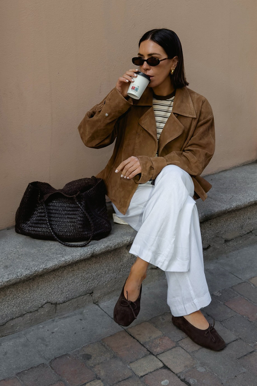 a woman wearing a tan suede jacket, white pants, brown flats, and woven brown leather bag