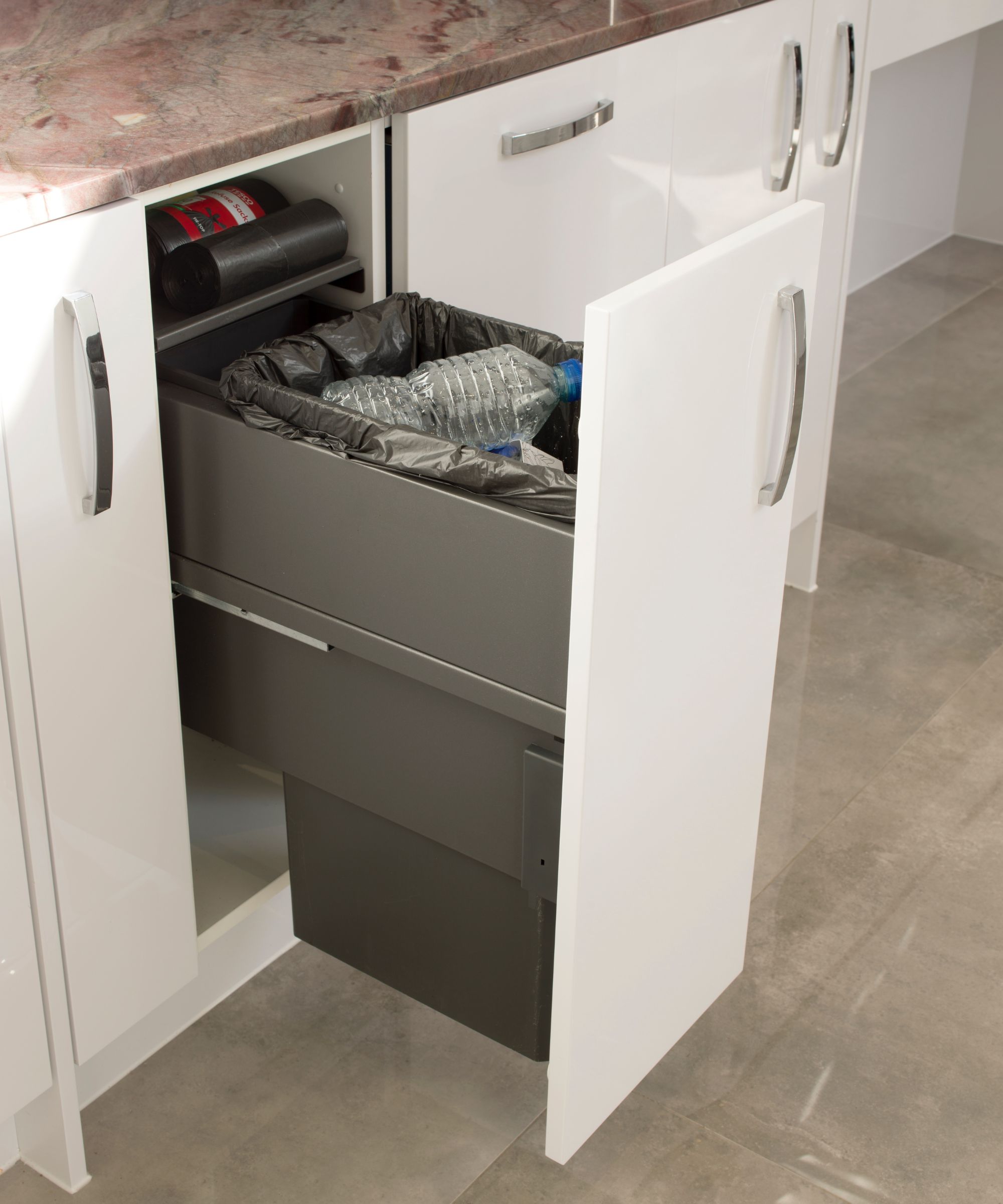 A pull-out kitchen bin integrated into a glossy white cabinet, with a black liner and compartments for recycling. A practical hidden waste storage solution beneath the worktop.