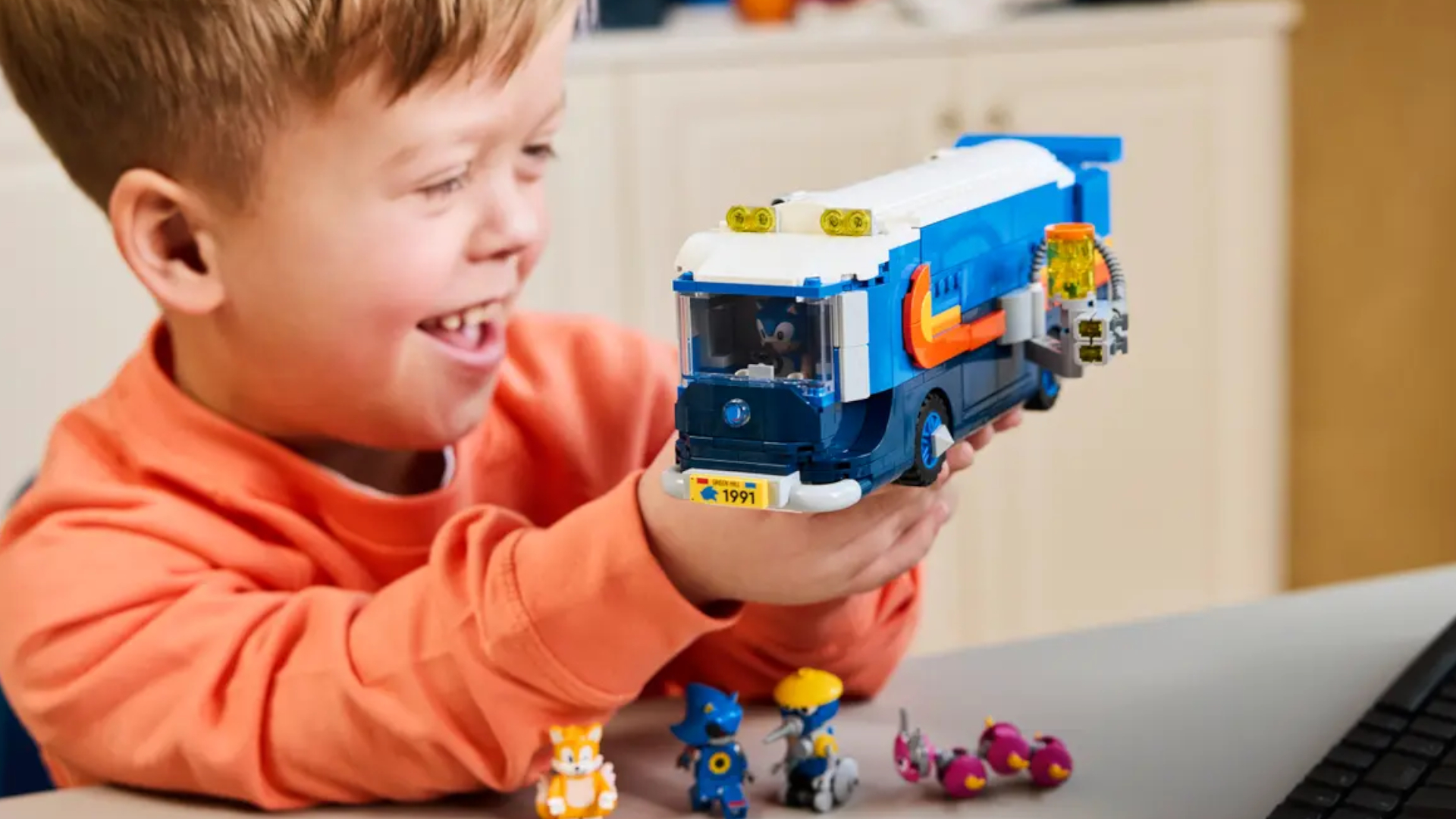 A smiling child holds up the Lego Team Sonic Command Truck, with minifigures on the gray surface beneath