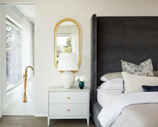 A bedroom with large black upholstered headboard, white bedside table, gold oval mirror and ensuite bathroom
