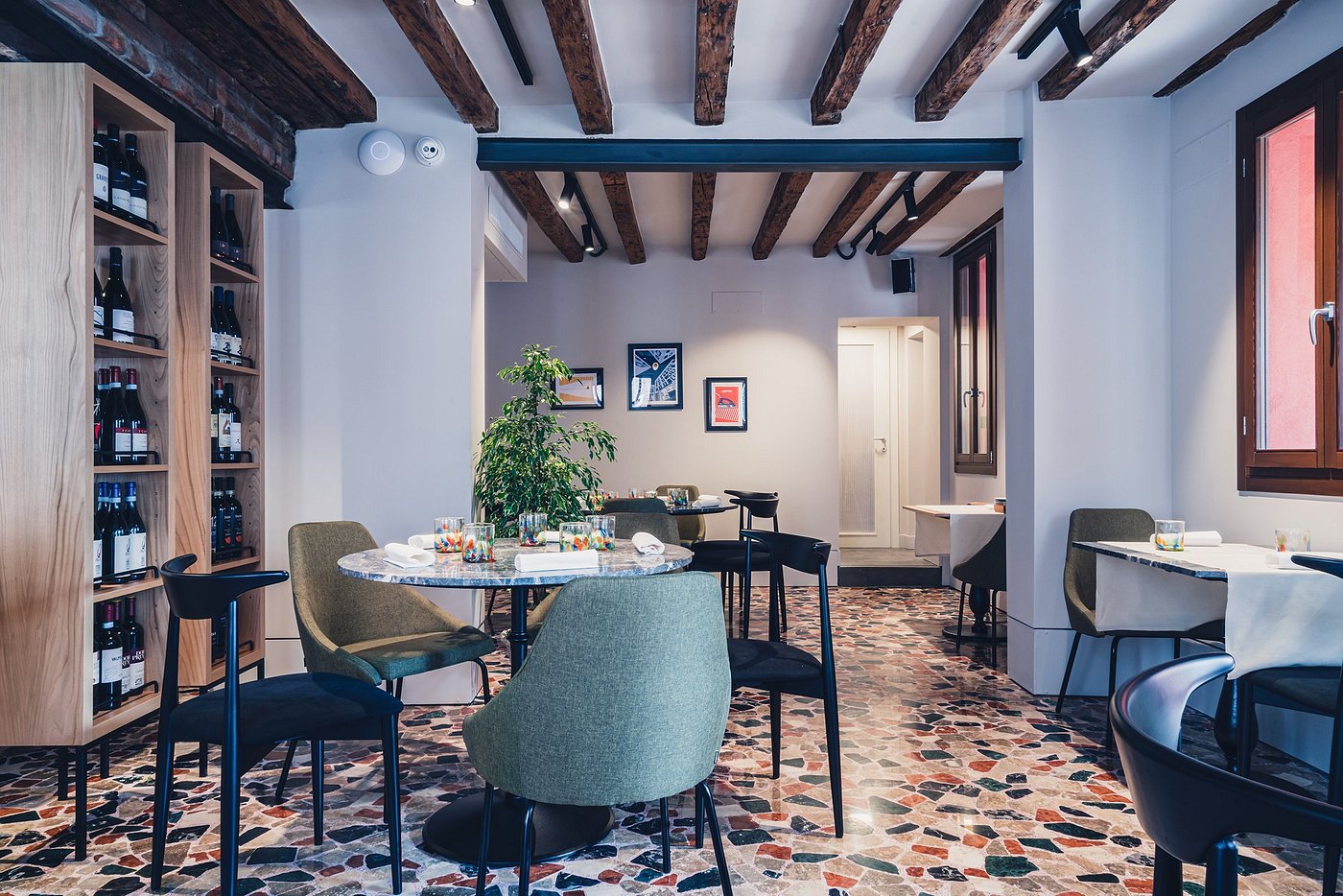 A warmly lit restaurant boasts terrazzo flooring in terracotta, light beige, and black hues, sculptural mid-century chairs, marble tables in round and square formats, exposed ceiling beams, and wooden shelves.