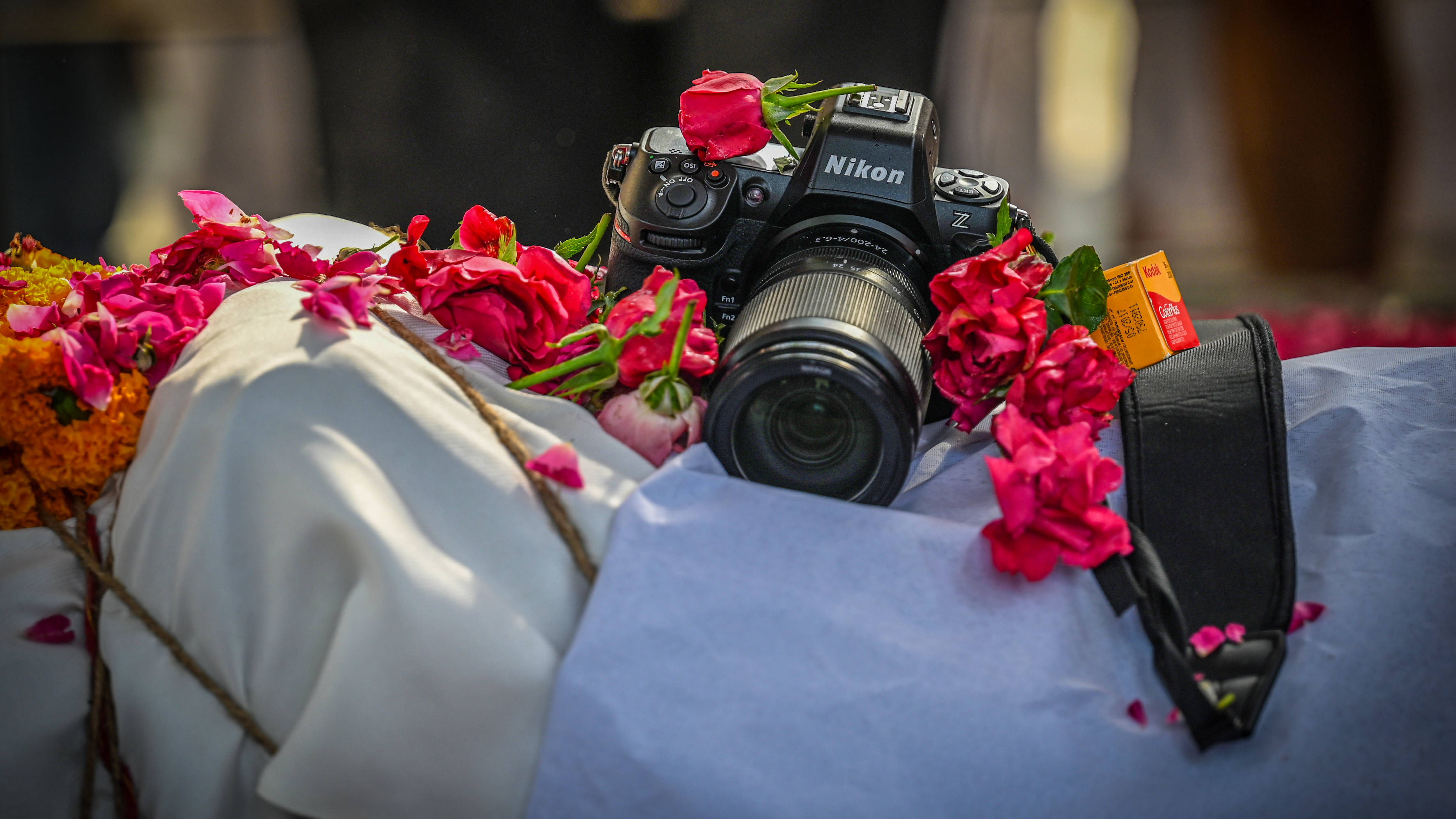 A professional Nikon mirrorless camera is surrounded by vibrant red and orange flowers atop a white and light blue cloth, with a small yellow Kodak film box tucked beside its strap.