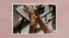 A close-up of a woman rubbing cream into her hands, whilst wearing a pink linen robe. A stylist wooden console table is also visible in the background, with beauty products on top/ in a pink watercolour paint-style template