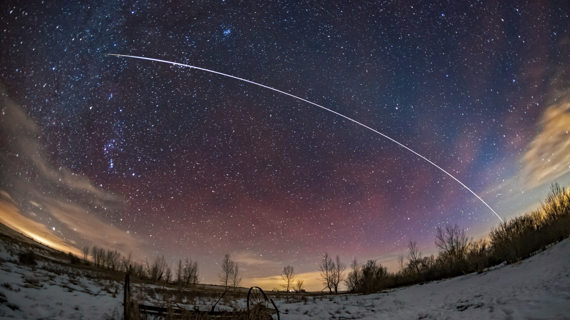 A timelapse photo showing the bright trail of the International Space Station leaving an arc in the night sky above a snowy landscape.