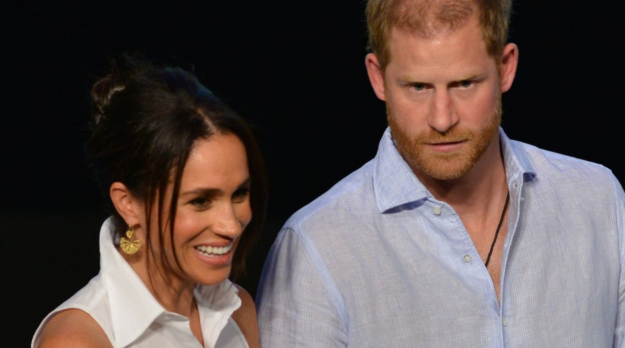 Meghan Markle wears a white, sleeveless shirt and gold earrings and wears her hair in a sleek updo, while Prince Harry wears a light blue shirt