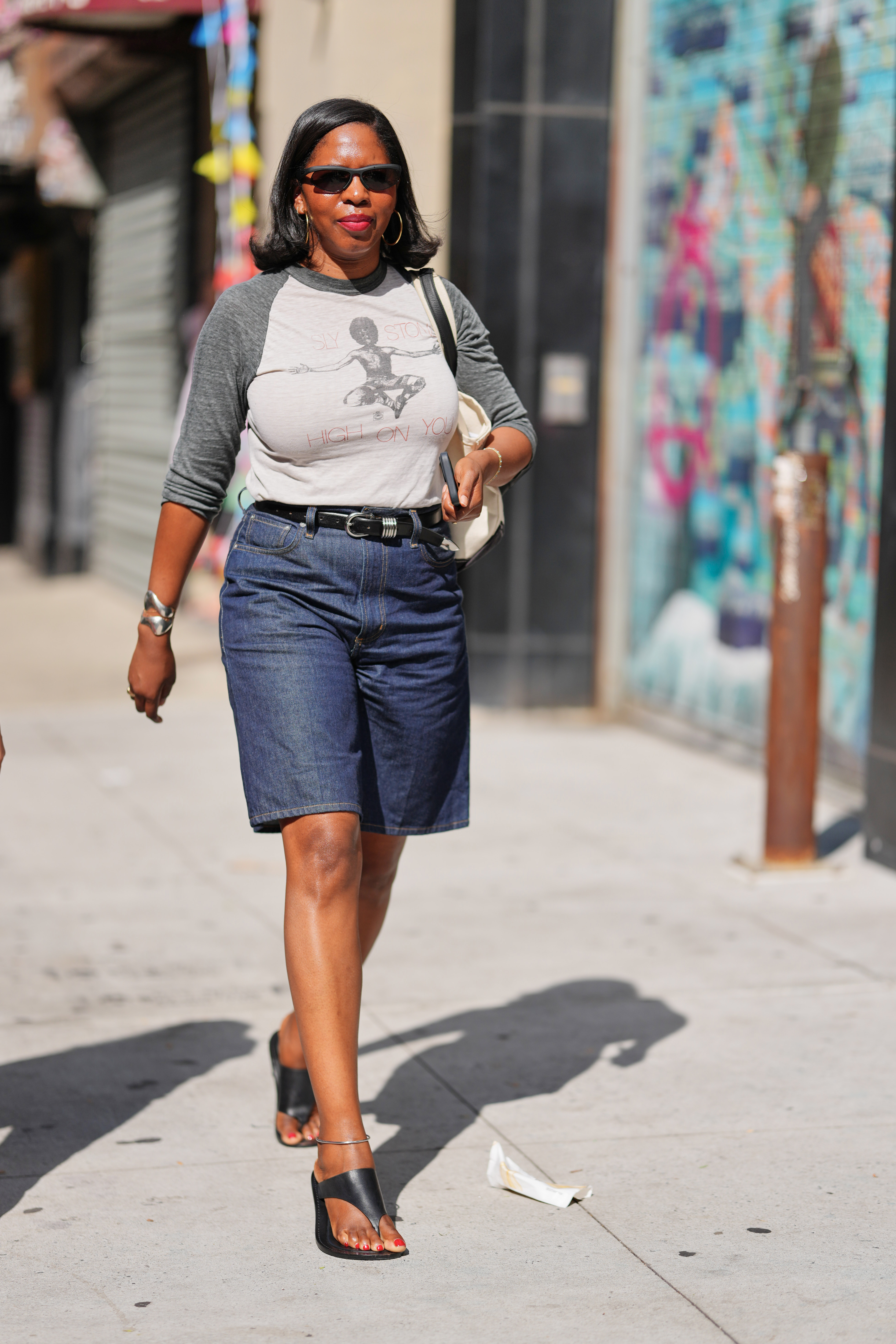 NEW YORK, NEW YORK - SEPTEMBER 12: A guest wears shoulder-length straight black hair parted at the side, slim black rectangular sunglasses, gold hoop earrings, a silver cuff bracelet on the left wrist and a thin gold bracelet on the right wrist with a gold ring on the left middle finger, a cream canvas tote bag with black shoulder straps, a three-quarter sleeve raglan baseball tee with a light gray body and dark gray sleeves featuring a faded black graphic and red printed text, high-waisted dark blue denim knee-length Bermuda shorts cinched with a black leather belt with a silver rectangular buckle, black leather open-toe sandals with a toe loop and low block heel with a silver ankle buckle, outside Ashlyn, during New York Fashion Week, on September 12, 2025 in New York, New York (Photo by Edward Berthelot/Getty Images)