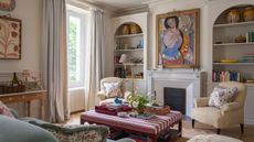 neutral London living room with a fireplace, alcove bookcases, large art, vintage objects, a striped ottoman, and two armchairs facing a blue couch