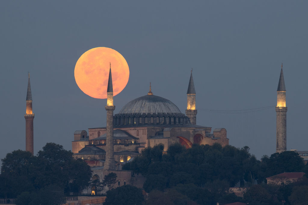 A lua cheia baixa no céu brilha em um tom laranja-rosado acima de uma grande mesquita.