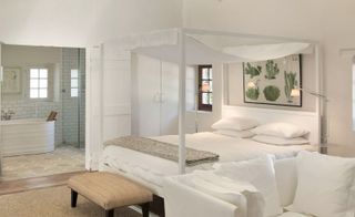 An interior look at a room at the Babylonstoren hotel. A large, white canopy bed sits on the right wall, with an image of the plants above it on the wall. To the far wall, we see a bathroom with white tiles and a bathtub.