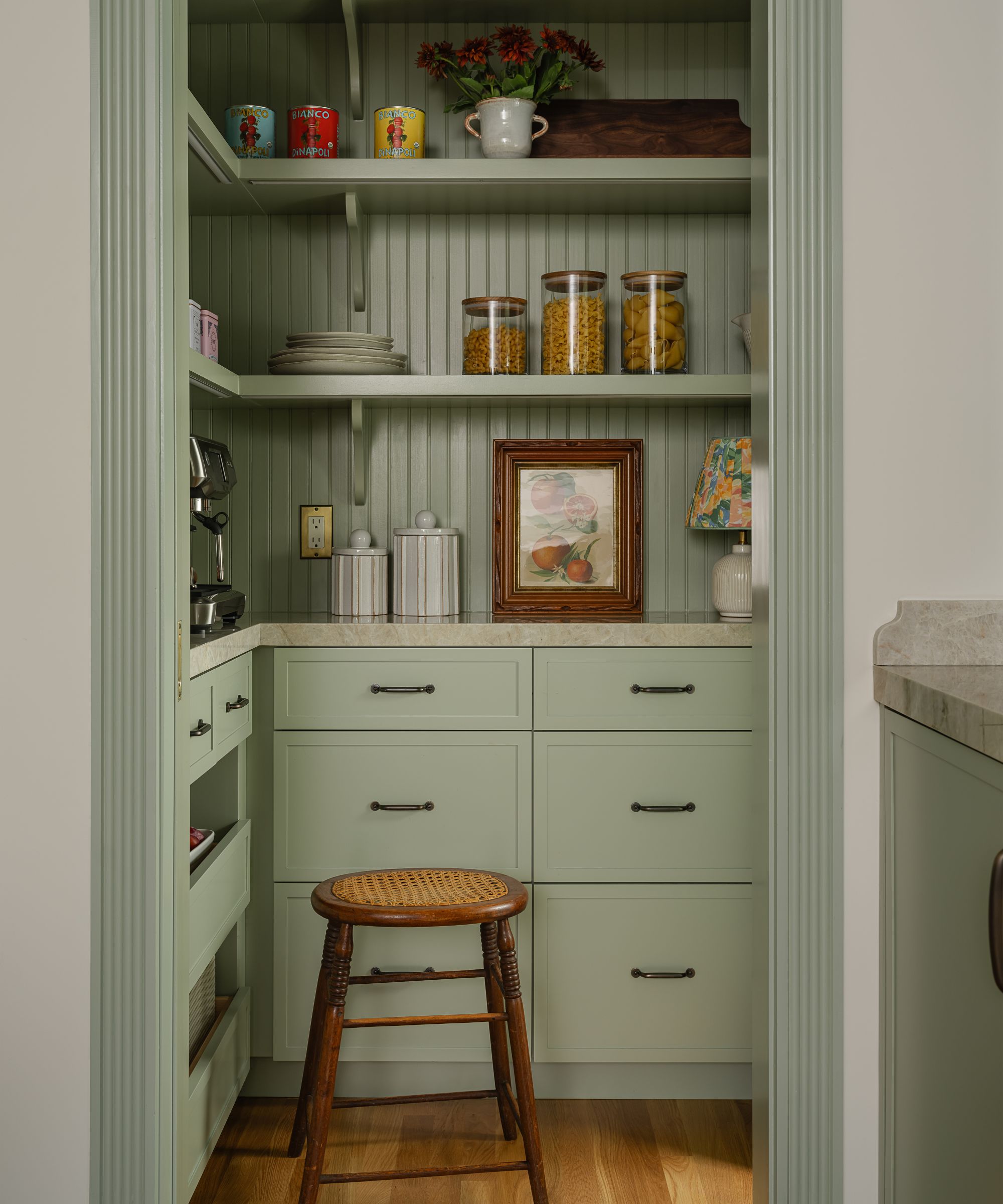 A close-up view of a walk-in pantry with sage green beadboard walls and matching cabinetry. The shelves are organized with glass jars of pasta, colorful canned goods, and a small framed botanical artwork