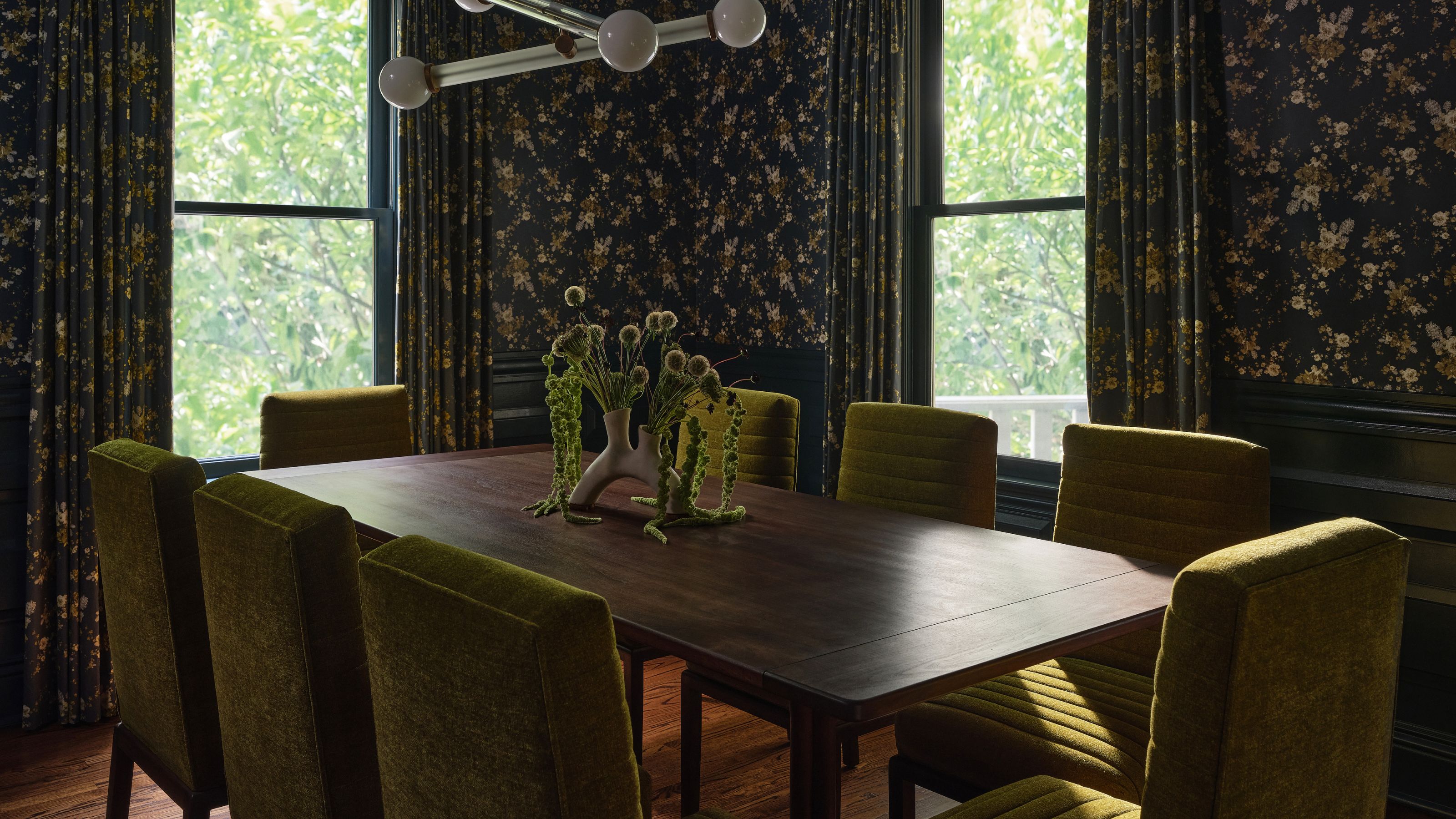 a dark green moody dining room with wooden table, velvet green chairs and wallpapered walls