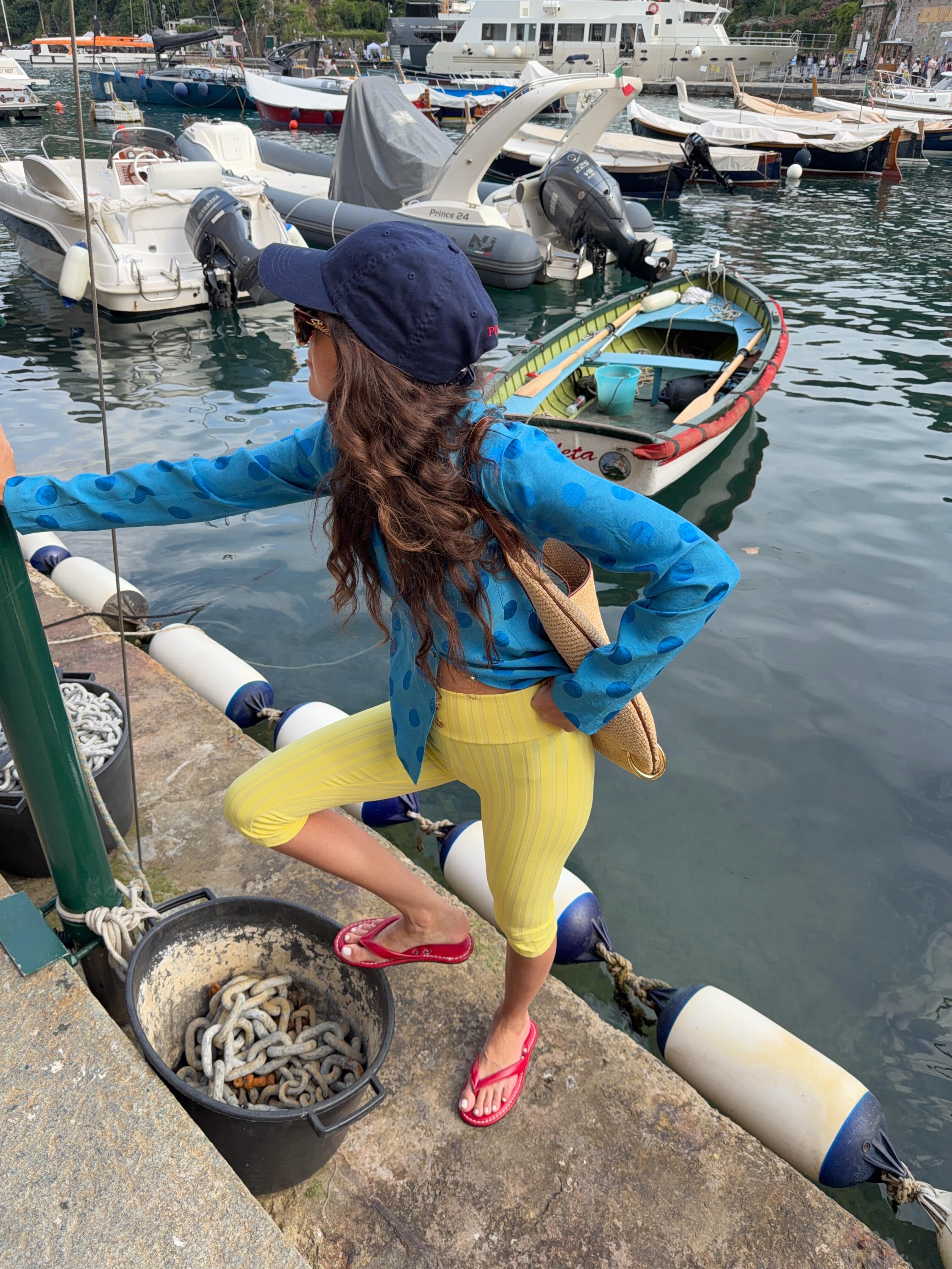 An image of an influencer wearing a navy baseball cap, blue button-down polka dot top, yellow capri leggings and red flip flops