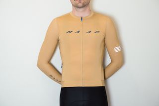 A white man in a tan long sleeve cycling jersey and black cycling shorts stands against a white wall