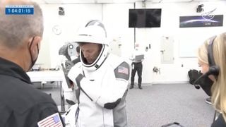 a man puts on a white flight suit in a large white room, with two technicians standing nearby to help.