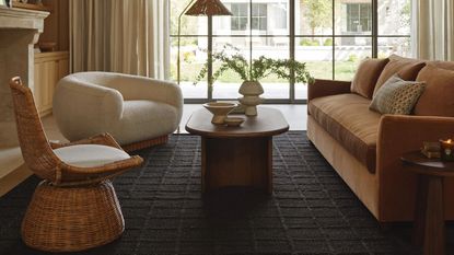 Living room with large windows, a dark rug, orange couch, cream armchair and rattan chair 