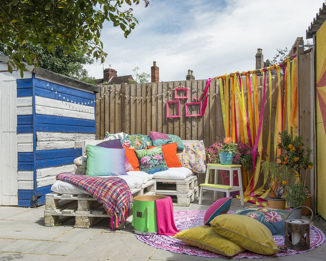 A colorful patio with a painted stroped shed and multi colored ribbon curtain along a fence with pallet furniture and a round rug