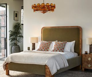 A plush bed frame, fitted with a mattress, sheets, and throw pillows, beneath a chandelier, against a white wall.