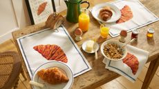 Image of a rustic wooden dining table with croissant placemats, blue and white plates with croissants on them, glasses of orange juice, toast, granola, and other breakfast items.