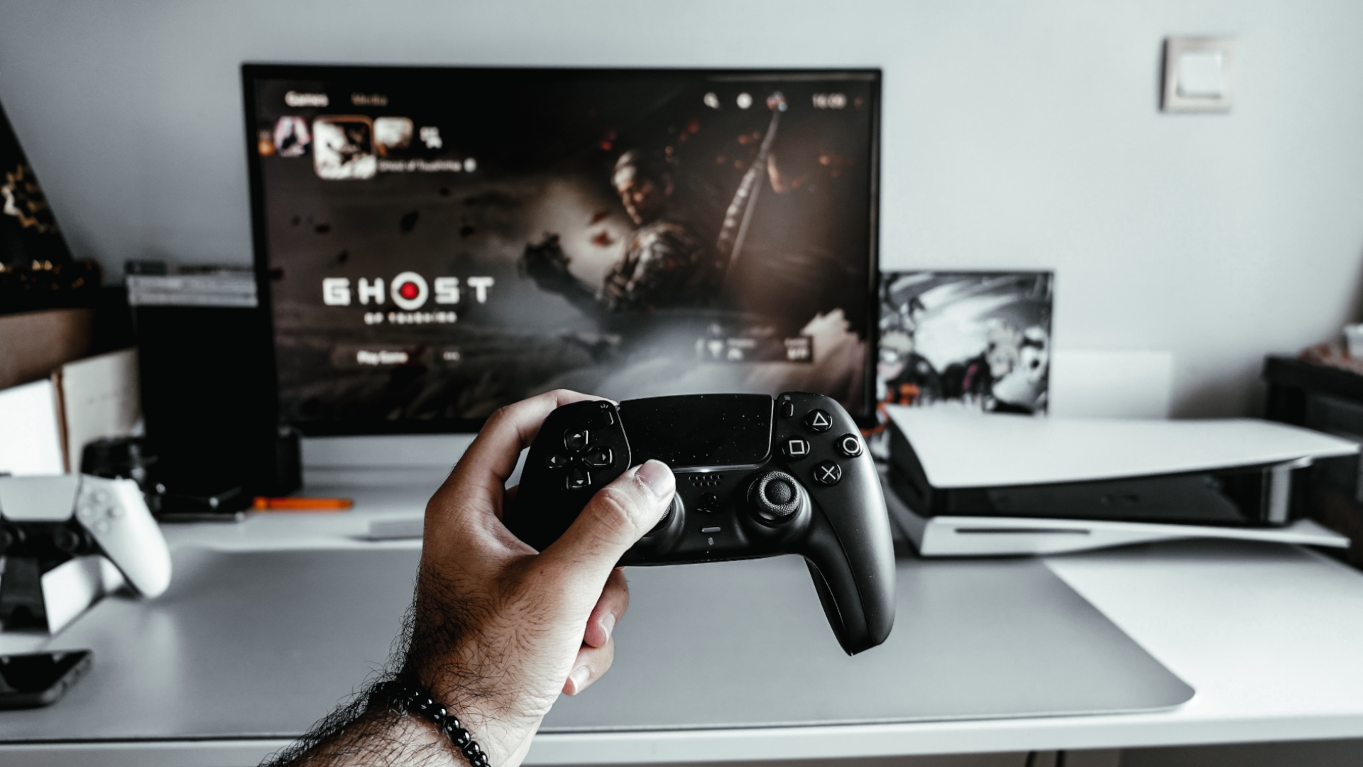 A man's hand holding a black PS5 DualSense controller in front of a TV showing Ghost of Tsushima