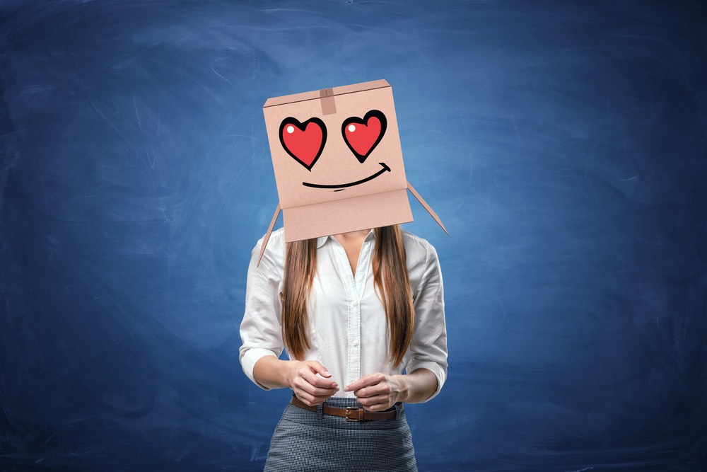 Young beautiful businesswoman with cardboard box on her head with drawn smiley face with hearts instead of eyes.