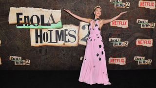 Millie Bobby Brown poses with arms outstretch in front of the Enola Holmes 2 logo at Netflix's NYC premiere on October 27, 2022.