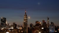 A photo of the New York skyline with the crescent moon shining next to the Empire State Building with a planet glowing toward the top of the screen. 