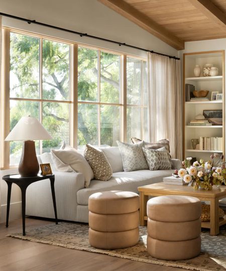 Modern neutral living room, two matching round ottomans, cream sofa, patterned rug, wooden coffee table, lamp on black side table, shelving in background with accessories