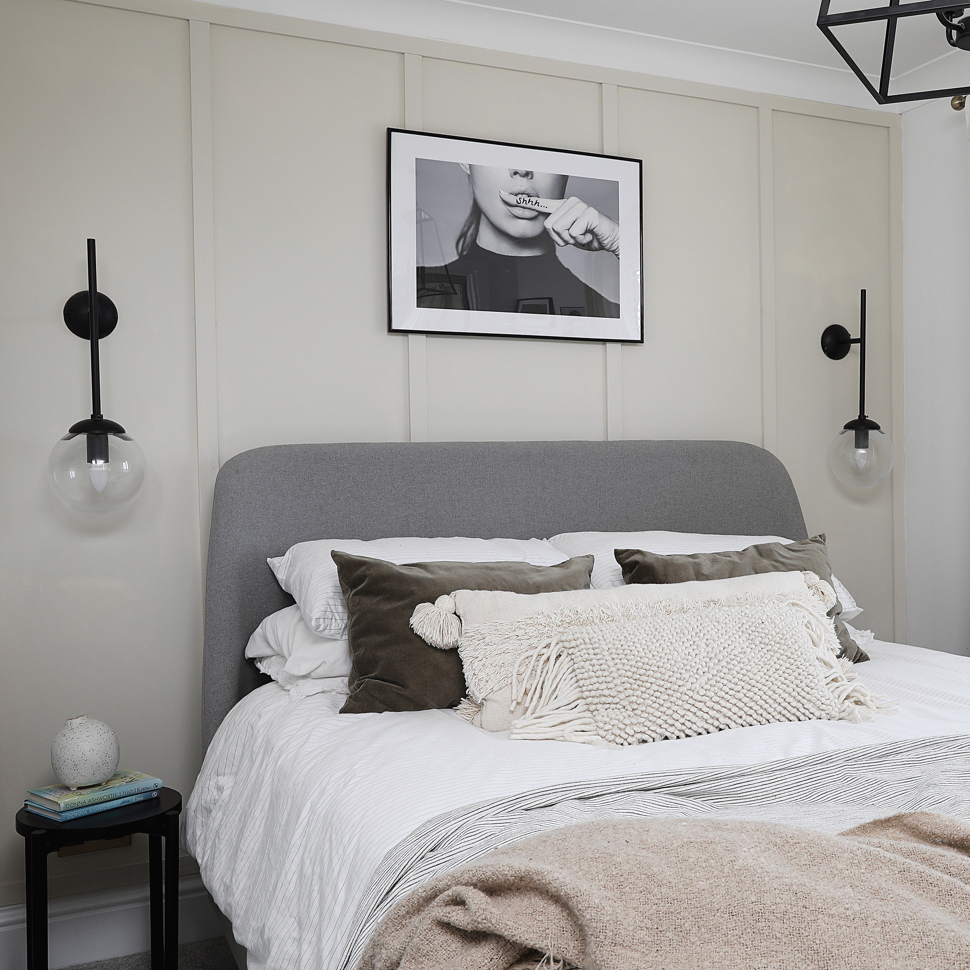 a neutral bedroom with beige panelled walls a grey upholstered headboard black globe wall lights black and white photo art