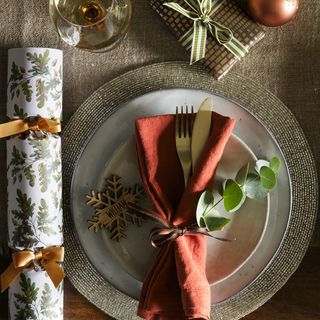 Christmas place setting with red napkin with cutlery on top of a gold rimmed plate, and a festive cracker next to it