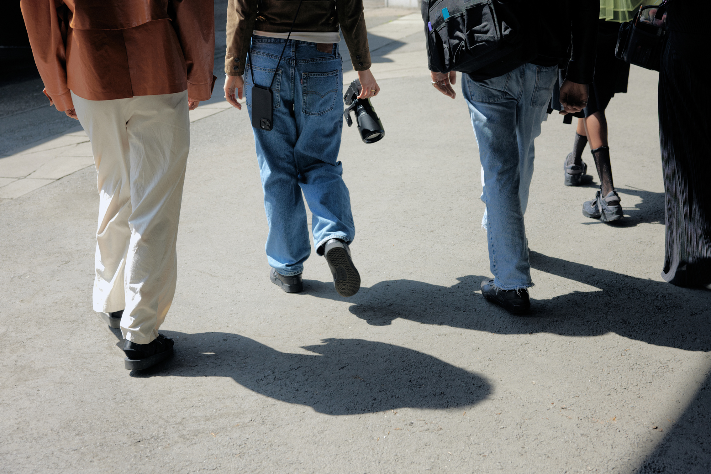 Four people walking on a sunlit pavement, one holding a camera, all wearing casual clothing and shoes