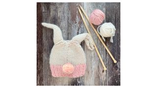A woollen knitted pink and white hat with bunny ears and a pink pom pom for a tail, next to some knitting needles and some white and pink balls of wool.