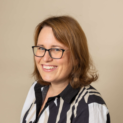 A professional headshot of a smiling woman with shoulder-length auburn hair and black-framed glasses, wearing a monochrome striped shirt against a neutral background.