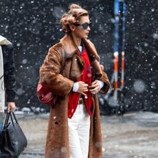 Bella Hadid wears white jeans with a red sweater, fur coat, and burgundy bag.