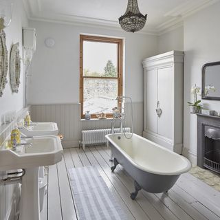 Large bathroom with central freestanding roll top bath and chandelier style pendant above. Painted loorboards and white walls