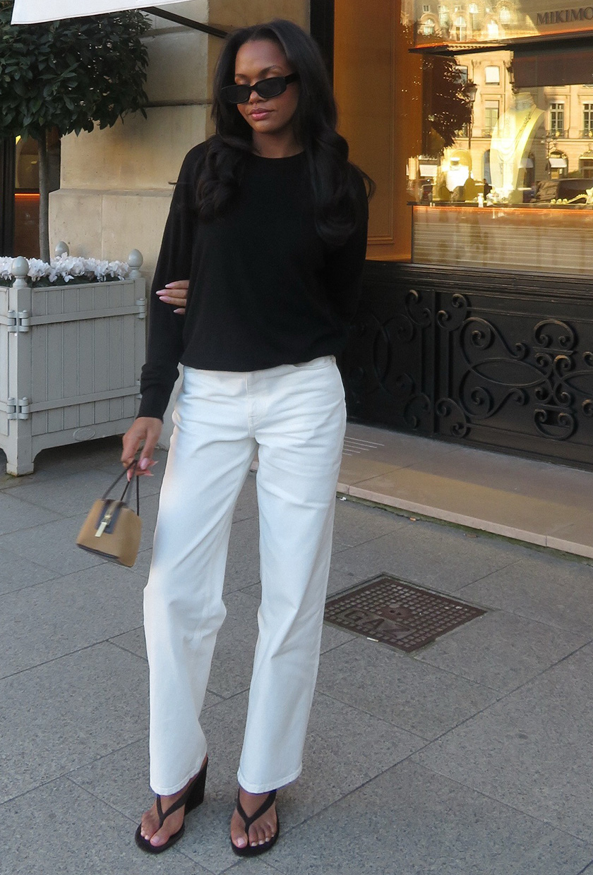 A woman in Paris wearing a black crewneck sweater with white jeans, a brown suede bag, thong wedge sandals, and black sunglasses.