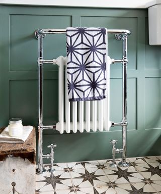Heated towel rail and radiator against a green wood panelled wall with black and white patterned tile floor