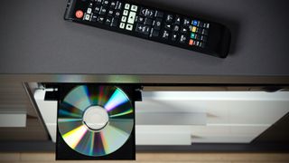 Blu-ray player on the top of a media console showing the disc being ejected and a black remote control. 