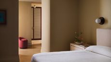 cream bedroom with cream bedding and headboard, red ottoman, wall sconce, and timber side table