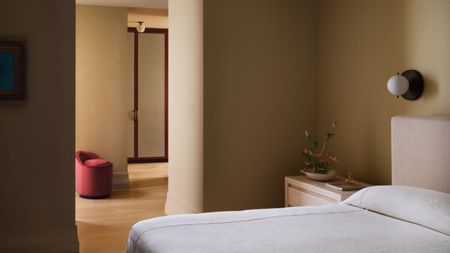 cream bedroom with cream bedding and headboard, red ottoman, wall sconce, and timber side table