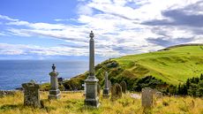 A tranquil coastal landscape featuring slender grave markers, rolling green hills, and a calm sea under a partly cloudy sky. Ideal for themes of history, memory, and nature.