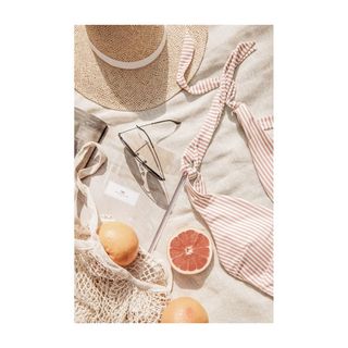 A warm-toned flatlay poster of beachy must-haves, including a netted tote bag, sliced grapefruit, a gingham swimsuit, and a straw hat
