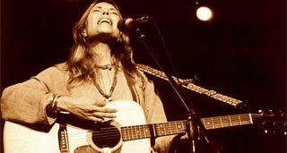 A classic sepia-toned live shot of Joni Mitchell playing her Martin dreadnought and singing live in 1974