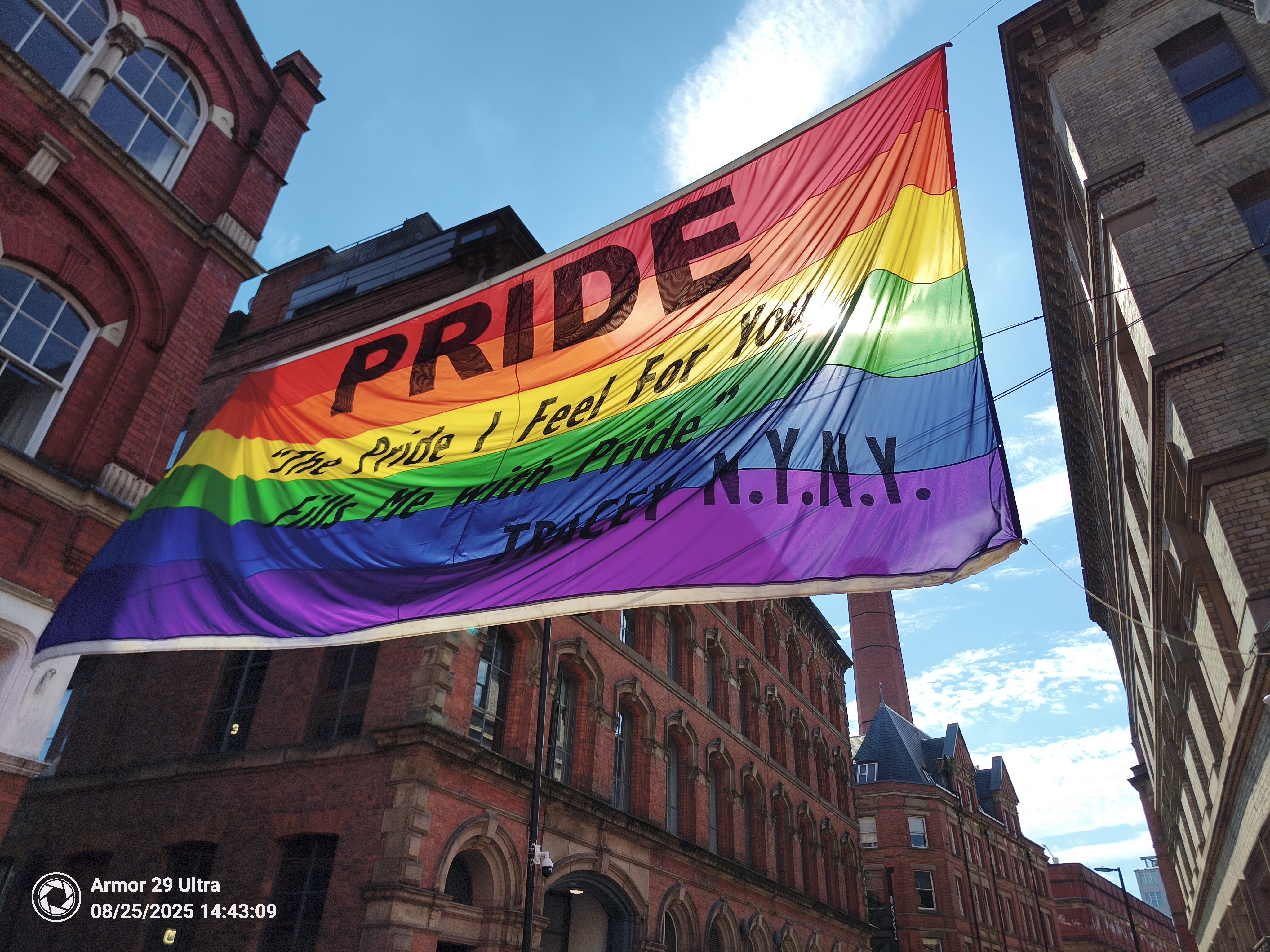 A huge rainbow-colored flag with the word "PRIDE" is hung between two brick buildings against a clear blue sky.