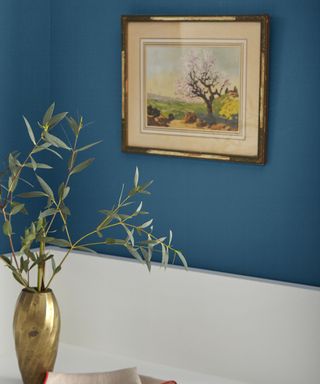 A bright blue wall with a framed painting and small vase in front